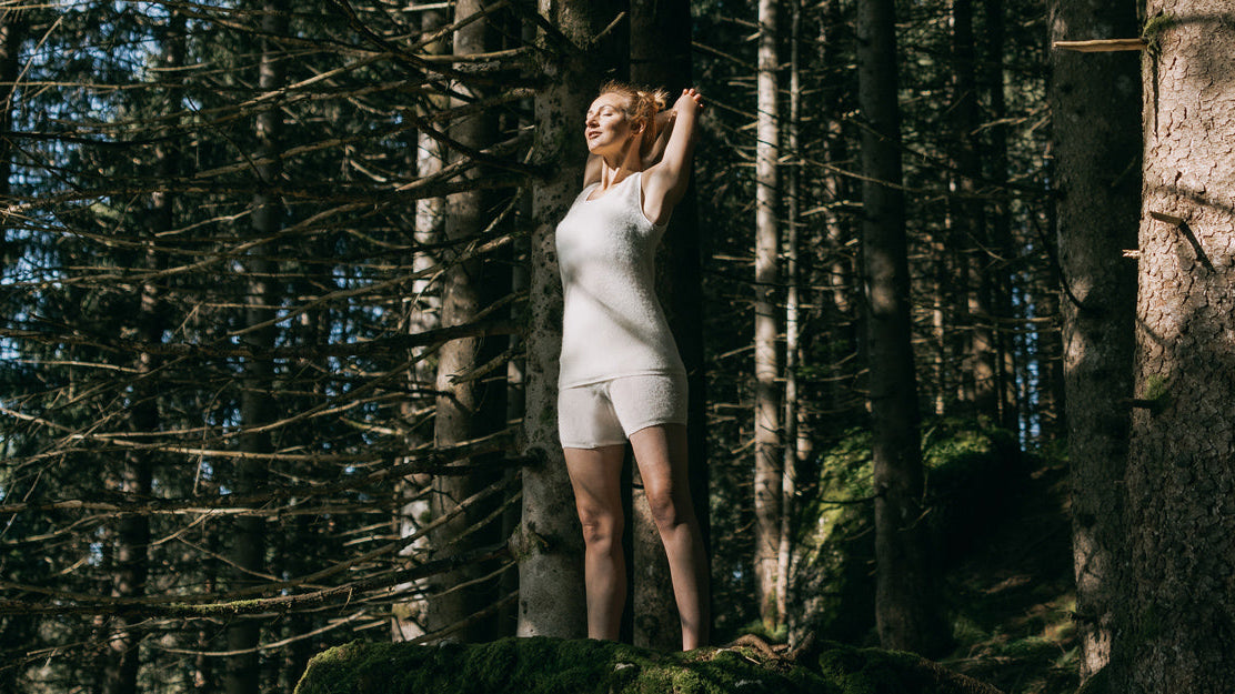 Eine junge Frau steht auf einem moosbedeckten Felsen in einem dichten Nadelwald, trägt ein weißes Tanktop und Shorts, Arme erhoben im Sonnenlicht.