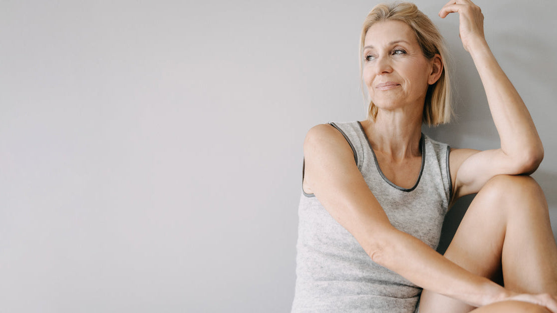 Eine mittelalte Frau mit kurzen blonden Haaren sitzt nachdenklich auf dem Boden, trägt ein graues ärmelloses Top und lehnt an einer Wand.