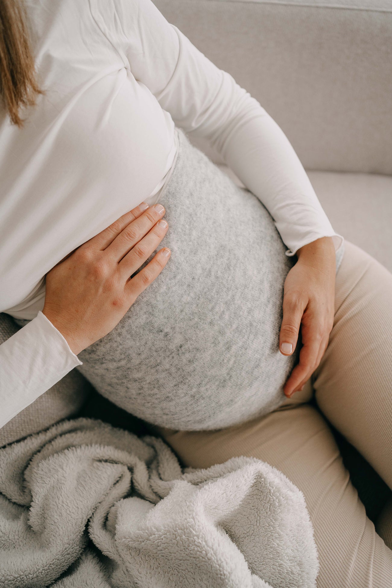 Person auf Sofa, hält sanft den Bauch. Trägt ALPAKA Rückenwärmer ALVA, der Wärme und Komfort bietet, ideal für empfindliche Haut und Wohlbefinden.