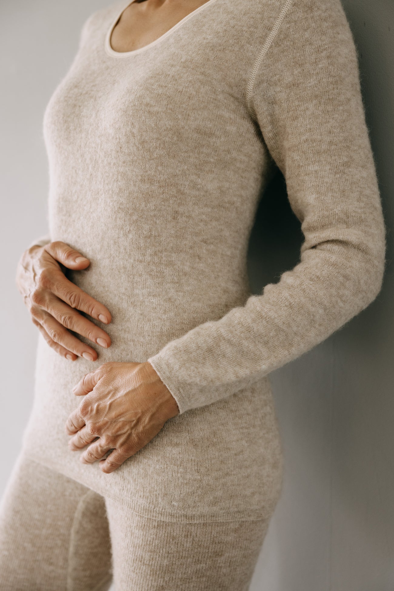 Person trägt ein ALPAKA Damen langarm Shirt AVELYNA aus weichem Strick, das nahtlos anliegt und Thermoeigenschaften bietet, während sie sich an eine Wand lehnt.
