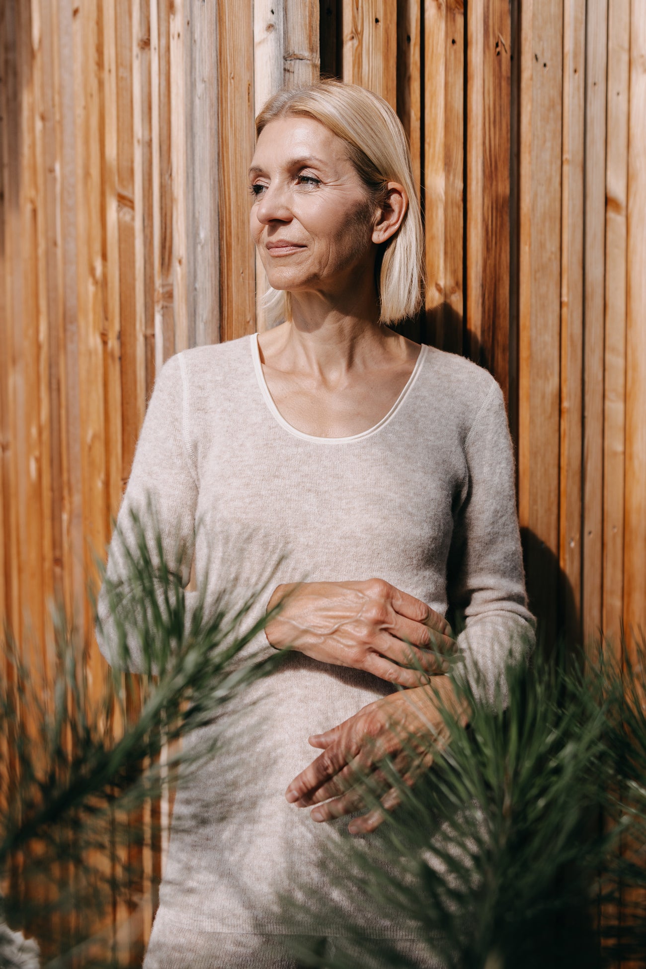 Frau trägt das ALPAKA Damen langarm Shirt AVELYNA, betont durch Sonnenlicht und umrahmt von unscharfen Zweigen, mit nachdenklichem Blick zur Seite.
