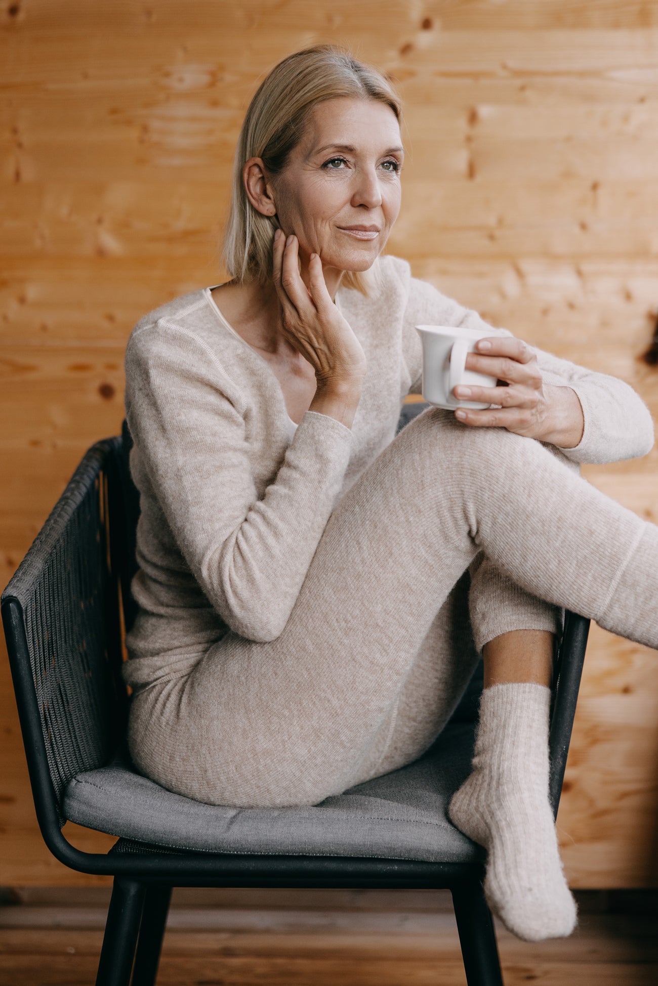 Eine Person trägt das ALPAKA Damen langarm Shirt AVELYNA aus beigem Strick, sitzt in einem Sessel und hält eine Tasse, wirkt entspannt und nachdenklich.