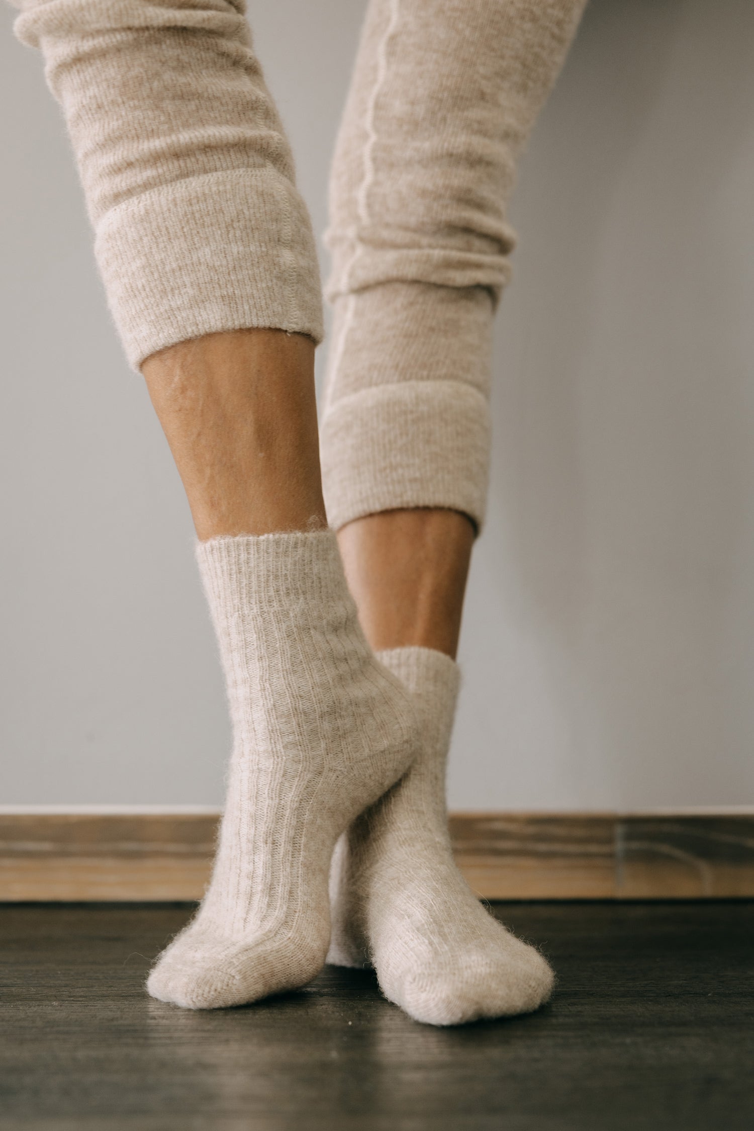 ALPAKA Kuschelsocken ALMA: Person trägt weiche, cremefarbene Alpakasocken auf dunklem Holzboden, bieten Wärme und Komfort durch atmungsaktive, flusenarme Alpakafasern.