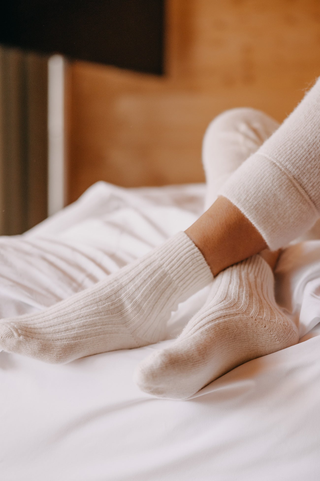 Person trägt ALPAKA Kuschelsocken ALMA, zeigt Beine in cremefarbenen, gerippten Strümpfen, auf einem Bett liegend. Socken bieten Wärme und Komfort durch Alpakafasern.