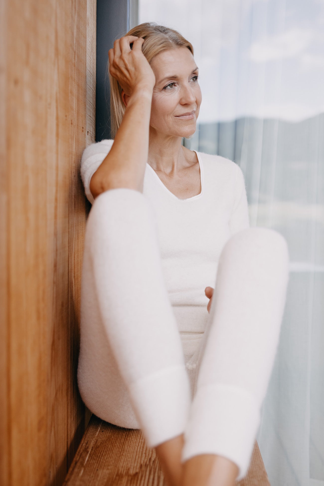 Frau in weißer ALPAKA Damen Leggins ALBERTA sitzt nachdenklich am Fenster, gekleidet in weiche Loungewear, verkörpert Wärme und Komfort.
