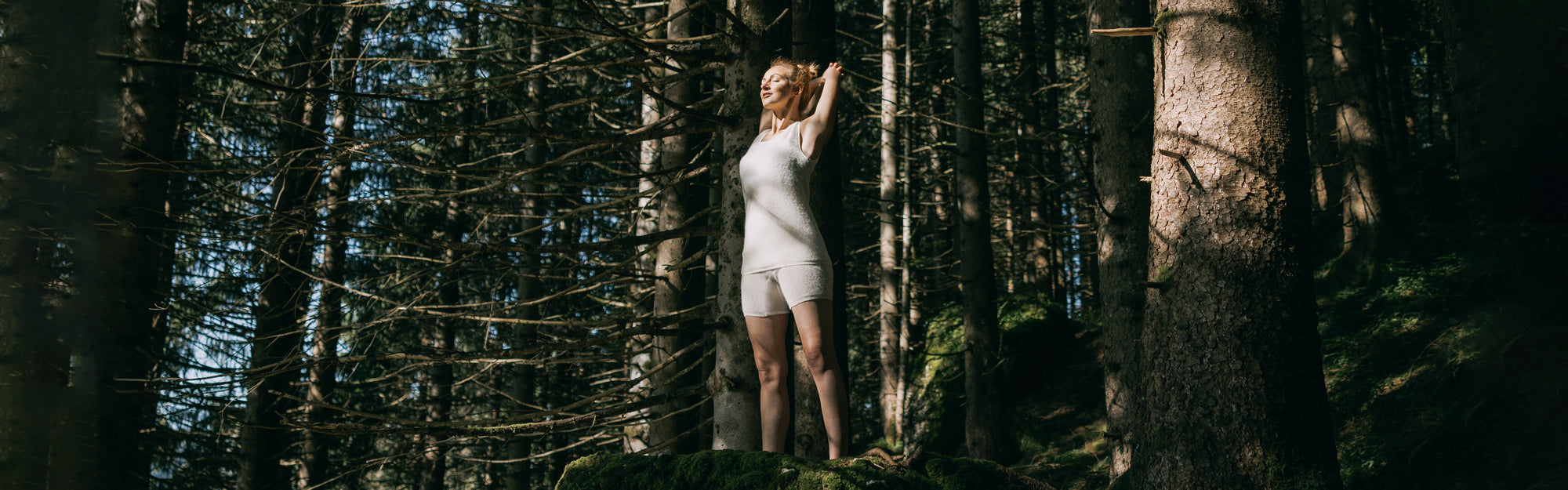 Eine junge Frau steht auf einem moosbedeckten Felsen in einem dichten Nadelwald, trägt ein weißes Tanktop und Shorts, Arme erhoben im Sonnenlicht.