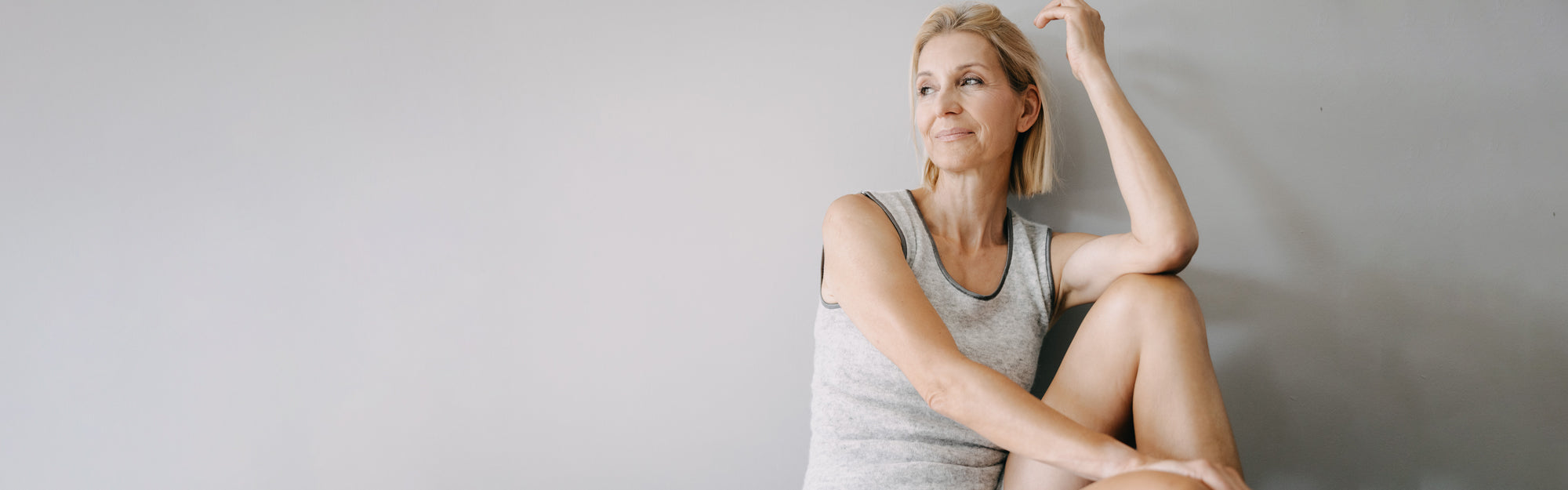 Eine mittelalte Frau mit kurzen blonden Haaren sitzt nachdenklich auf dem Boden, trägt ein graues ärmelloses Top und lehnt an einer Wand.