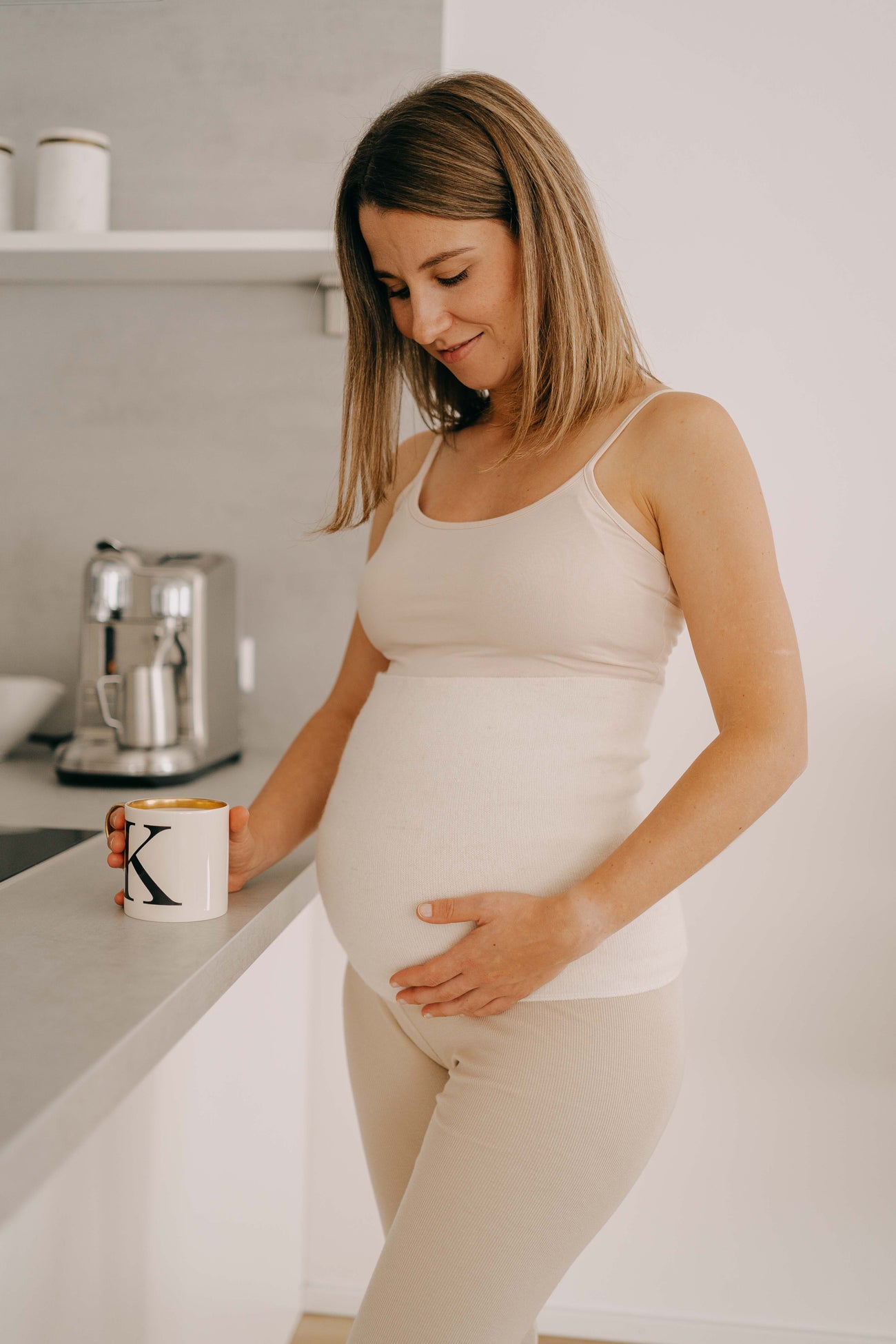 Schwangere Frau in minimalistischer Küche trägt ALPAKA Rückenwärmer ALVA, hält Babybauch, in der anderen Hand eine Tasse mit „K“. Espresso-Maschine im Hintergrund.