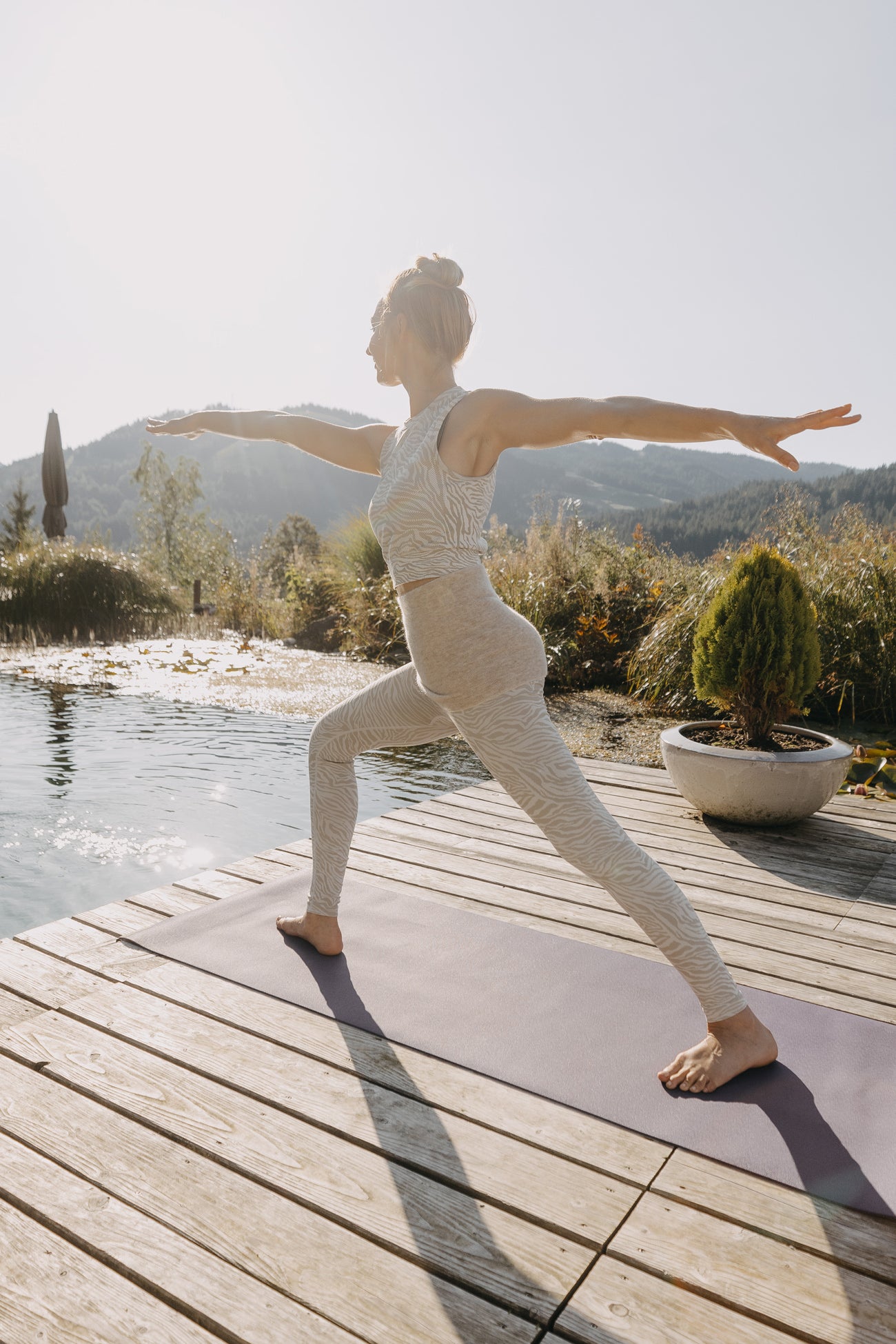 Frau in Warrior II-Pose auf lila Yogamatte; trägt ALPAKA Rückenwärmer ALVA für Komfort und Wärme. Holzdeck am Teich, umgeben von Natur.