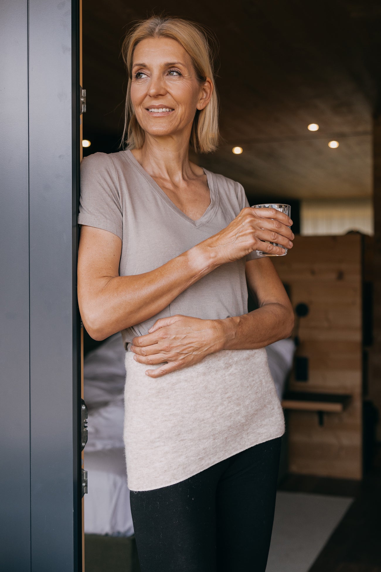 Frau lehnt entspannt an Türrahmen, trägt ALPAKA Rückenwärmer ALVA, der Komfort und Wärme bietet, ideal für empfindliche Haut und Verspannungen.