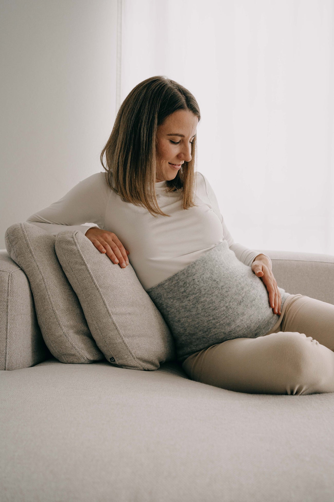 Schwangere Frau auf Sofa trägt ALPAKA Rückenwärmer ALVA, der Rückenwärme bietet. Sie lächelt sanft, genießt den Komfort in einem ruhigen Raum.