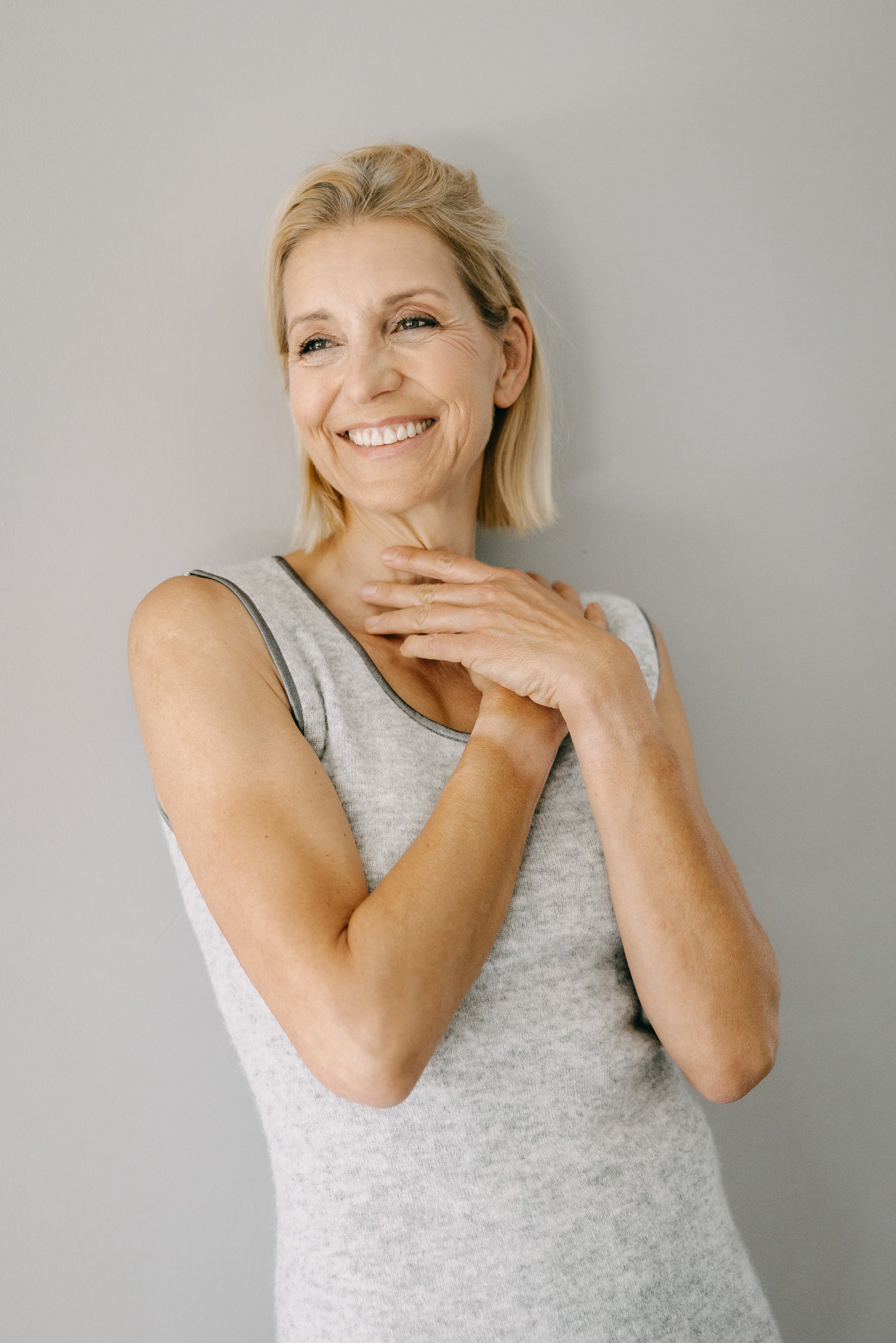 Mittlere Frau trägt ALPAKA Damen Top AYDA, lächelt sanft, Hände an der Brust. Top ist ärmellos, hellgrau, betont Weichheit und Thermoeigenschaften der Alpakafaser.
