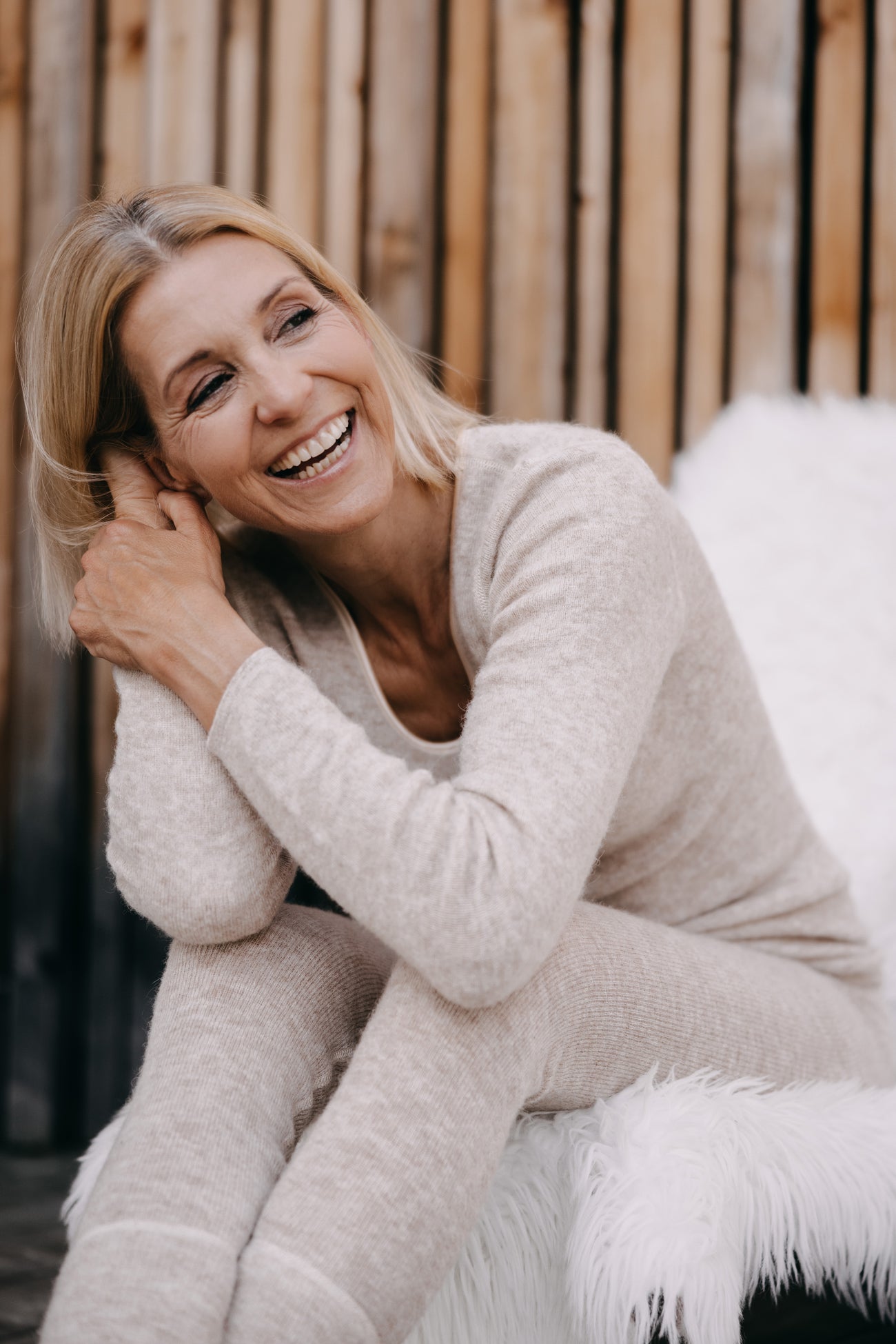 Frau in beigem ALPAKA Damen langarm Shirt AVELYNA lacht, sitzt auf weißer Decke; betont luxuriöse Weichheit und atmungsaktive, selbstreinigende Eigenschaften.