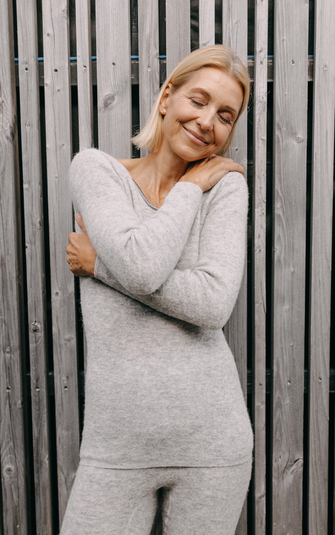 Frau in grauem ALPAKA Damen langarm Shirt AVELYNA lächelt mit geschlossenen Augen, Arme verschränkt, zeigt Komfort und Weichheit des Kleidungsstücks.