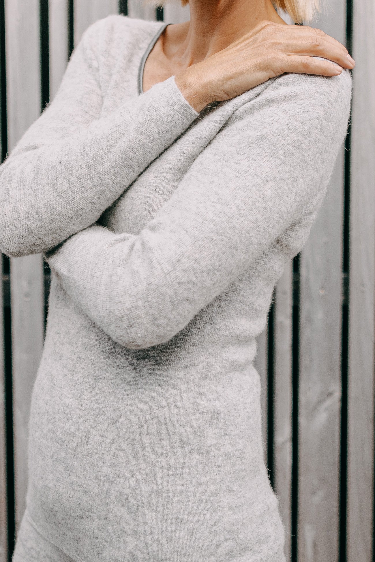 Person trägt ein ALPAKA Damen langarm Shirt AVELYNA aus hellgrauem Strick, Hand auf Schulter. Das Shirt bietet Komfort und Temperaturausgleich durch nahtlose Alpakafaser.