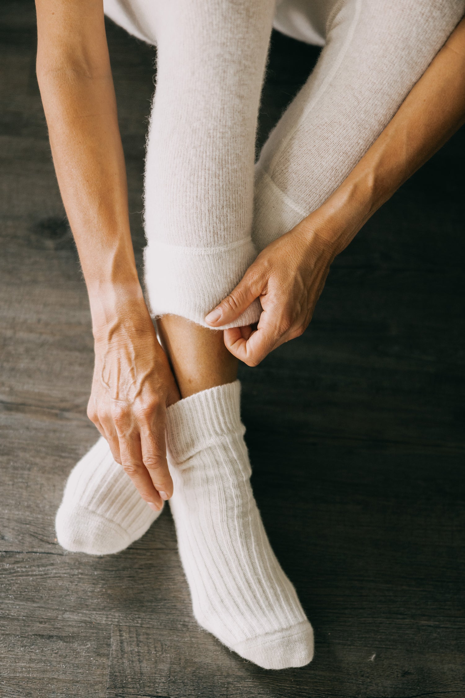 Nahaufnahme von Beinen, die ALPAKA Kuschelsocken ALMA anziehen, zeigt die gerippte, cremefarbene, gestrickte Struktur, die Wärme und Komfort bietet.