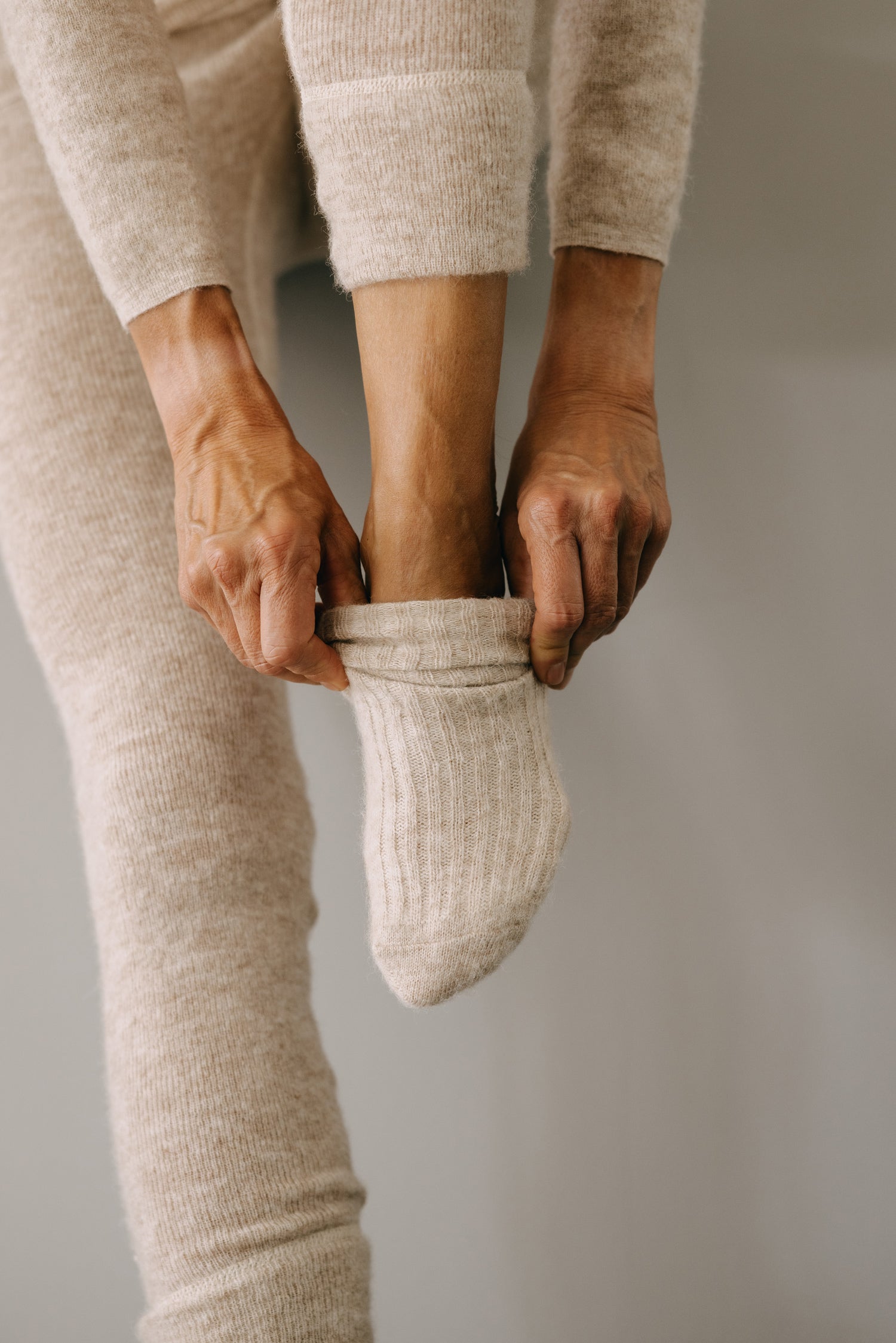 Nahaufnahme einer Hand, die einen dicken, gerippten ALPAKA Kuschelsocken ALMA über den Fuß zieht, getragen mit passenden beigen Strickleggings für optimalen Komfort und Wärme.