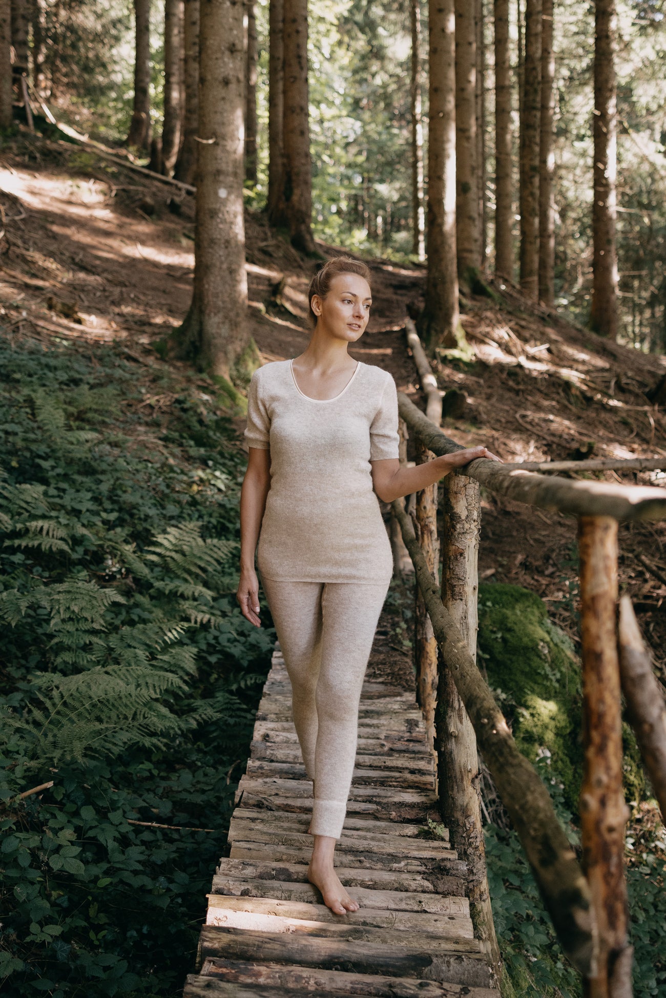 Junge Frau trägt ALPAKA Damen Leggins ALBERTA, geht barfuß über einen rustikalen Holzsteg im Wald, umgeben von Bäumen und moosigem Boden in sanftem Sonnenlicht.